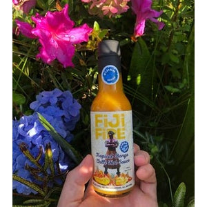 Hand holding Fiji Fire Tropical Bongo Chilli Hot Sauce bottle in garden with flowers